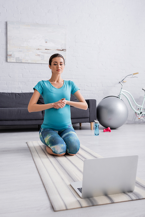 young pregnant woman in sportswear exercising near laptop in living room