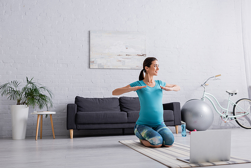 happy pregnant woman in sportswear exercising near laptop in living room