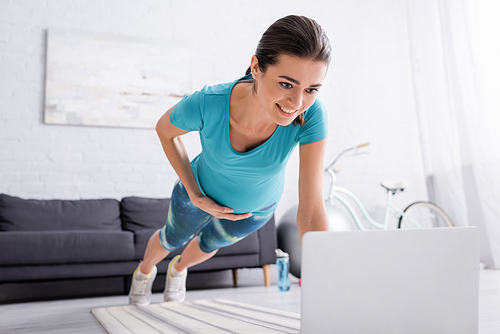 happy pregnant woman with sportswear working out near laptop in living room