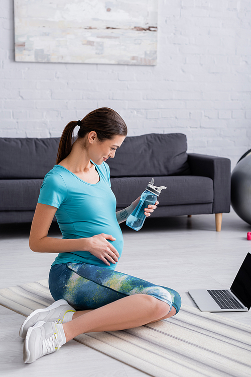 smiling pregnant woman looking at laptop while holding sports bottle