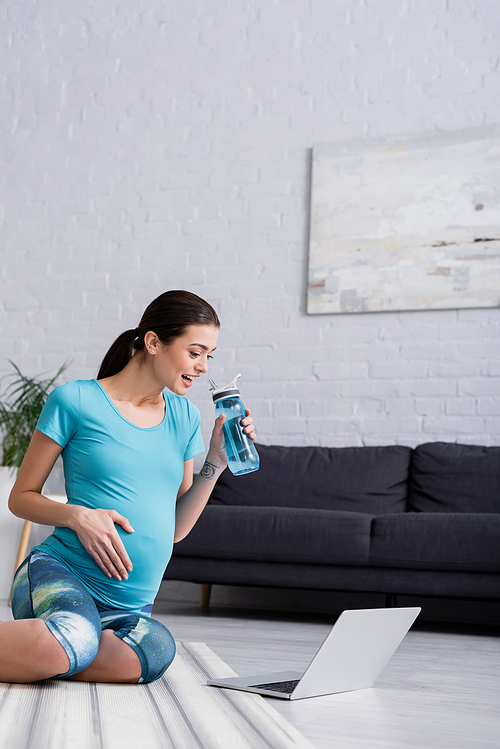 excited pregnant woman looking at laptop while holding sports bottle
