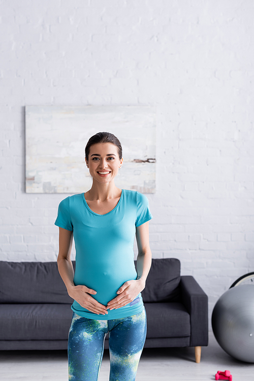 positive pregnant sportswoman touching belly in living room