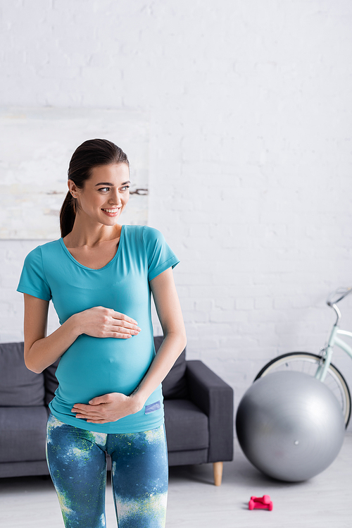 pleased young pregnant sportswoman touching belly in living room