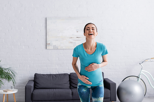 cheerful pregnant sportswoman touching belly and laughing in living room