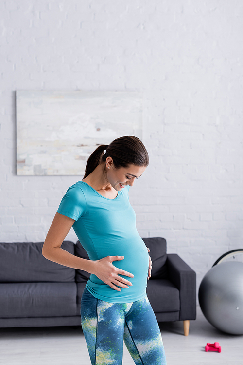 cheerful pregnant sportswoman touching belly in living room