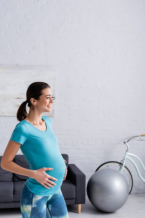 joyful pregnant sportswoman touching belly in living room