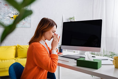 sick freelancer sneezing in paper napkin while working near monitor with blank screen