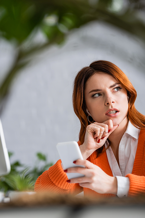 pensive woman holding smartphone and looking away while working at home on blurred foreground