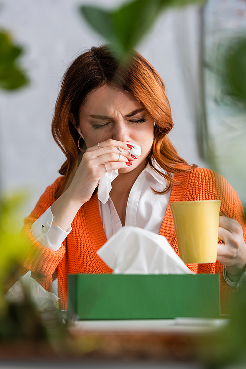 sick woman with closed eyes sneezing in paper napkin while holding cup of warm tea on blurred foreground