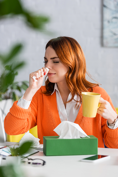 sick woman with closed eyes sneezing in paper napkin while sitting at home workplace with cup of tea