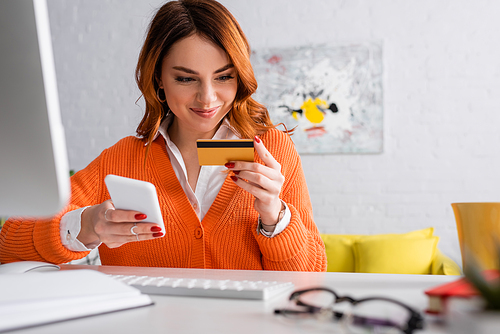 smiling woman holding credit card and smartphone while working at home