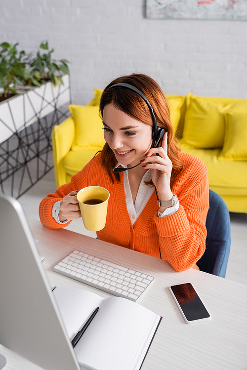 smiling woman in headset holding cup of tea while working near blurred computer at home