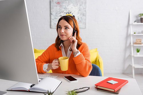 smiling woman in headset holding cup of tea while working near computer and smartphone with blank screen