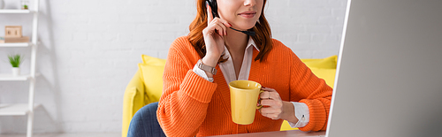 cropped view of woman in headset holding cup of tea while working near computer monitor, banner