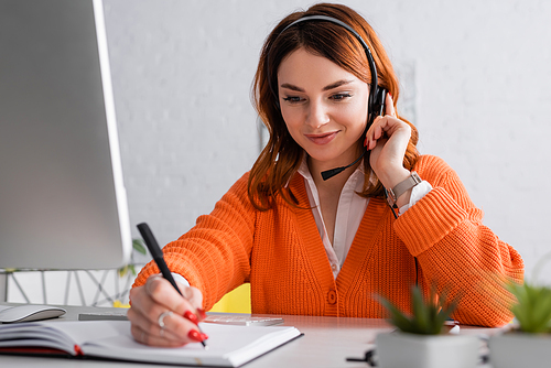 smiling woman in headset writing in notebook while working at home