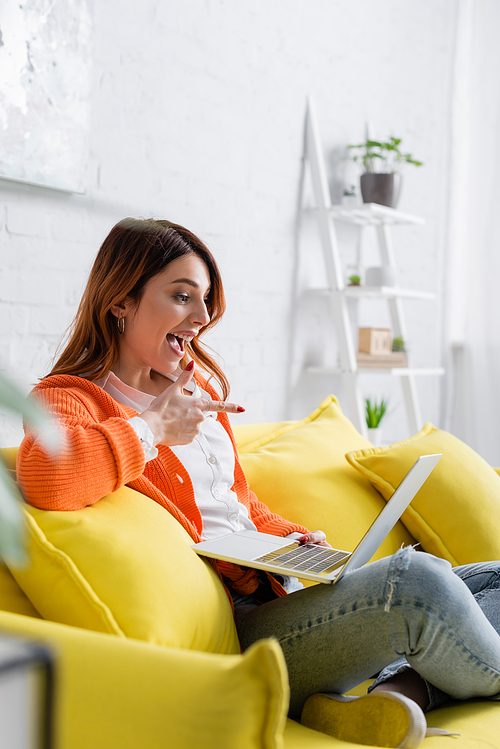 amazed freelancer pointing at laptop while sitting on yellow couch on blurred foreground