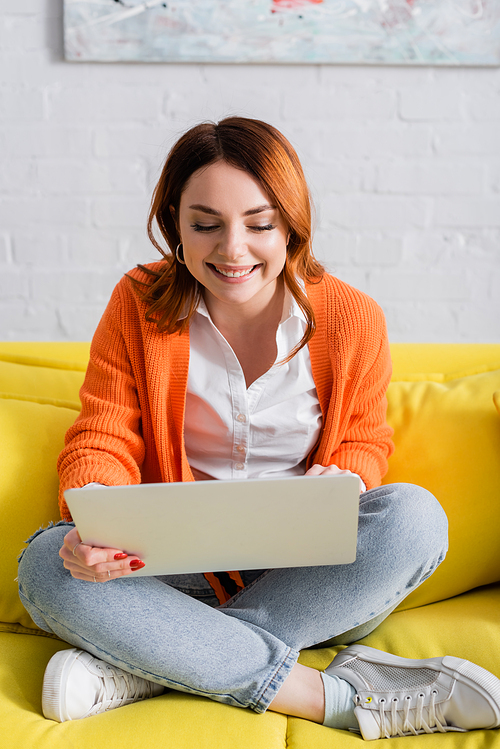 cheerful freelancer working on laptop while sitting on yellow sofa with crossed legs