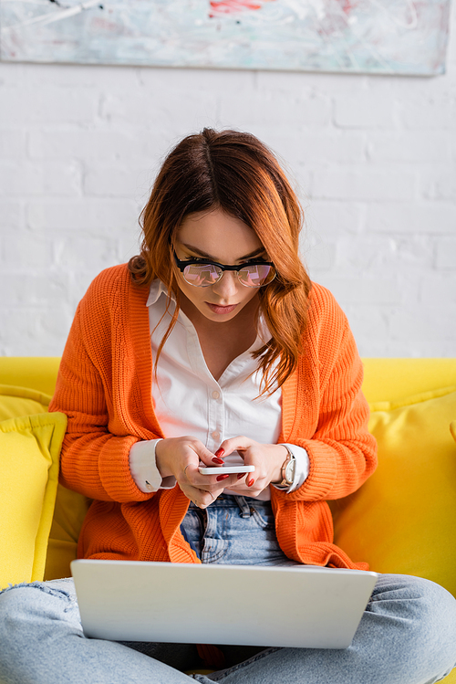 young woman in eyeglasses using smartphone while sitting with laptop on yellow couch