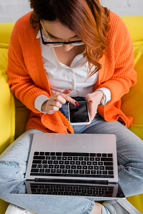 high angle view of woman in eyeglasses sitting on couch with laptop and using smartphone with blank screen