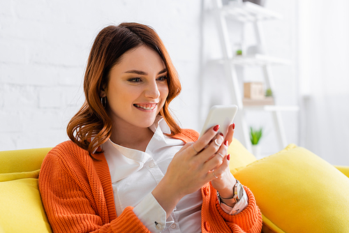 excited woman sitting on yellow couch and messaging on mobile phone