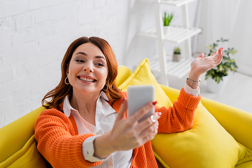 happy young woman sitting on yellow sofa and taking selfie on mobile phone