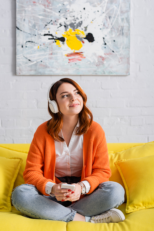 happy woman with smartphone sitting on yellow couch and listening music in headphones