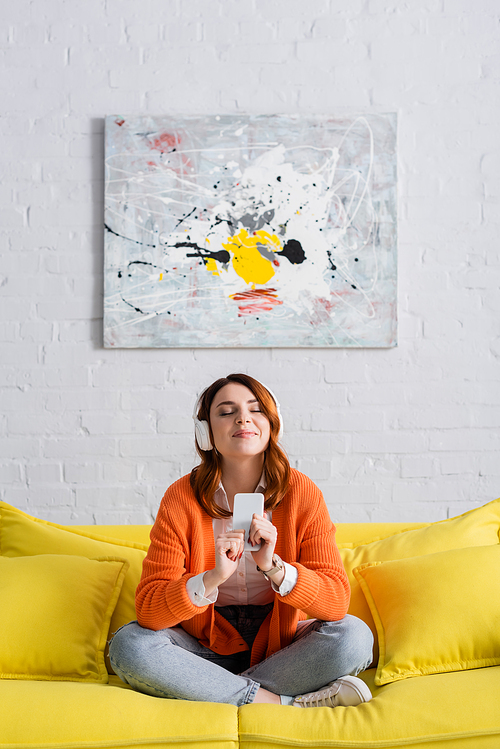 pleased woman with closed eyes listening music while sitting on yellow couch with crossed legs