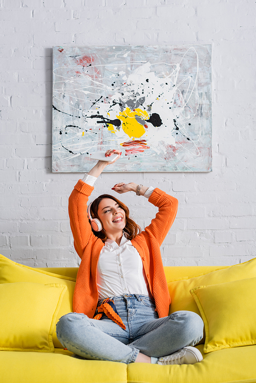 cheerful woman sitting on couch with raised hands while listening music in headphones