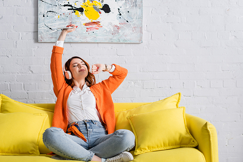 excited woman with smartphone in raised hand listening music on yellow couch