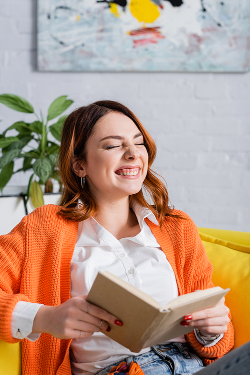 cheerful woman laughing with closed eyes while sitting on couch with book