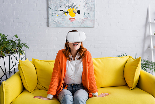 amazed woman in vr headset sitting on yellow couch in living room