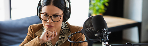 pensive asian radio host in glasses and headphones near microphone with pop filter during podcast, banner