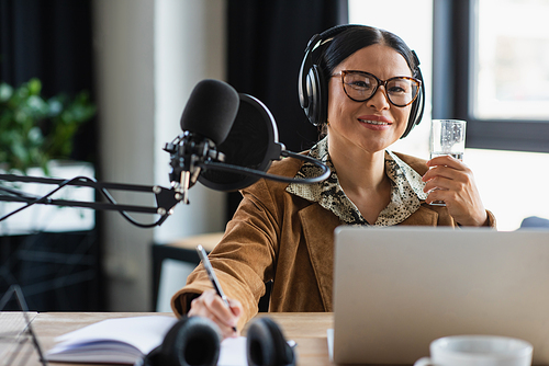 positive asian radio host in headphones holding glass of water and pen near notebook in studio