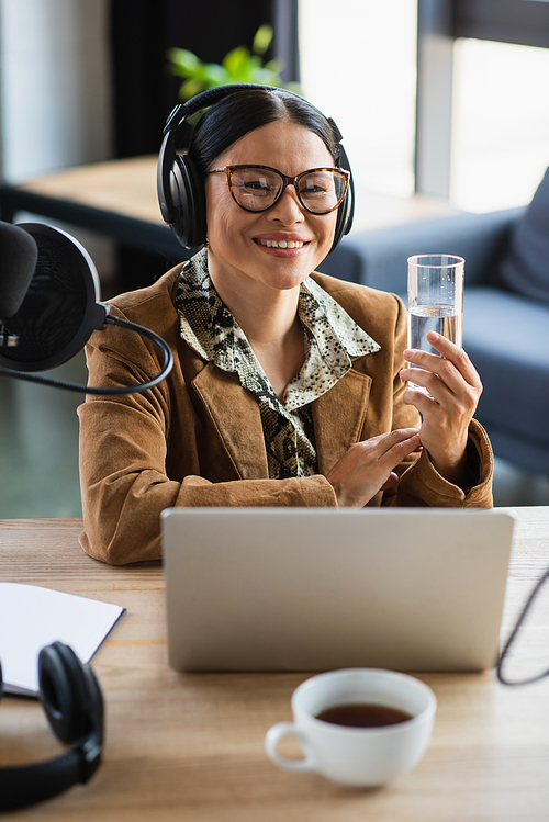 cheerful asian radio host in headphones holding glass of water near laptop in studio