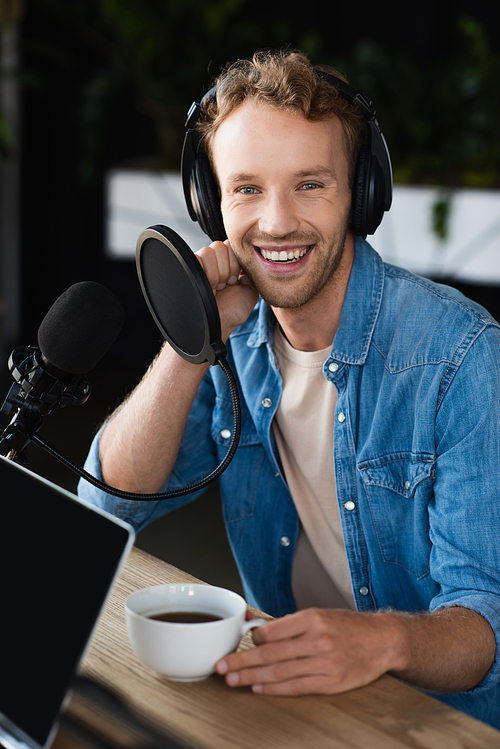 positive radio host in headphones holding cup of coffee in podcast studio