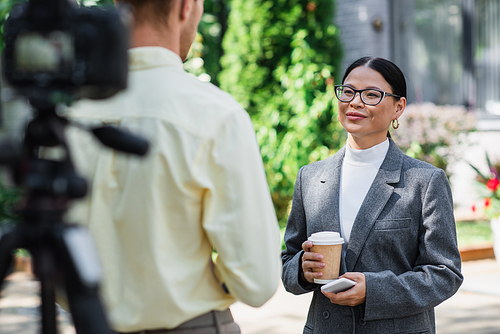 cheerful asian businesswoman in glasses holding smartphone and paper cup near blurred reporter