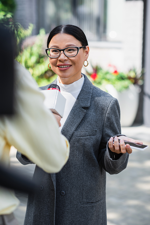 cheerful asian businesswoman in glasses holding smartphone and giving interview near blurred reporter