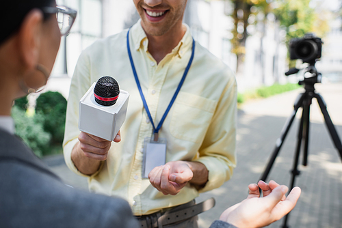 cheerful reporter holding microphone near blurred businesswoman gesturing while giving interview