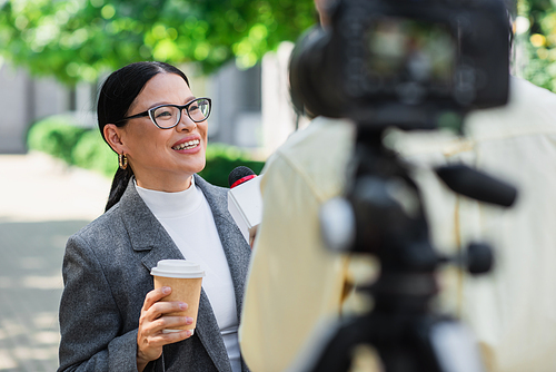 blurred reporter holding microphone near happy asian businesswoman with paper cup giving interview