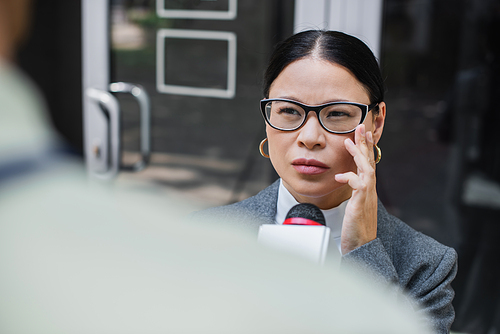 blurred reporter with microphone near pensive asian businesswoman adjusting glasses