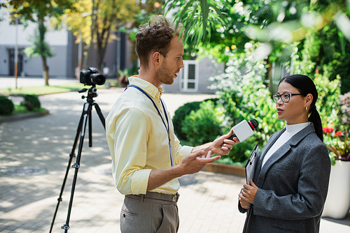 journalist gesturing while holding microphone and taking interview of asian businesswoman in glasses