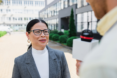 positive asian businesswoman in glasses looking at blurred journalist