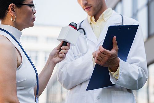 brunette asian journalist holding microphone near doctor in white coat with clipboard