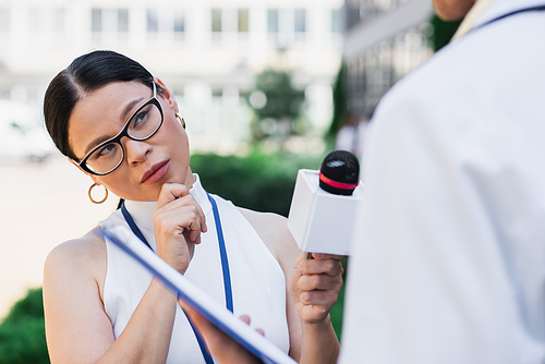 brunette asian journalist holding microphone and looking at blurred doctor in white coat