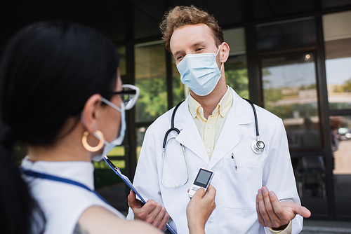 blurred asian reporter holding voice recorder near doctor in medical mask and white coat