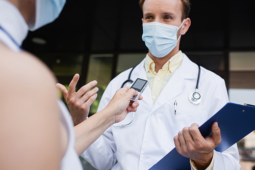 blurred reporter holding voice recorder near doctor in medical mask and white coat gesturing during interview