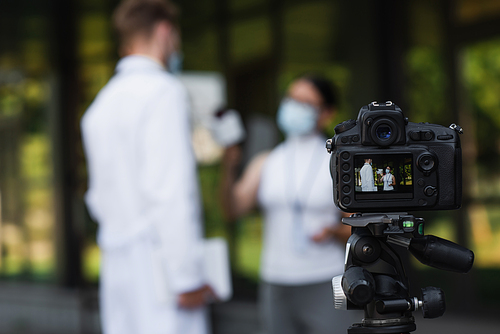 digital camera with asian journalist holding microphone near doctor in medical mask during interview on screen