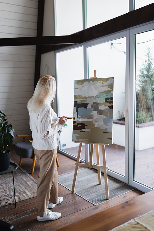 Back view of woman with paintbrush looking at canvas at home