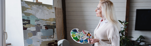 Side view of senior woman holding palette and brush near painting, banner