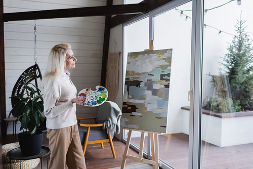 Side view of senior woman looking at canvas on easel in living room
